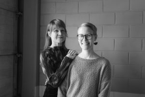 Elisa Itkonen ja Suvi Koivisto seisovat tiiliseinän edustalla, hymyilevät.