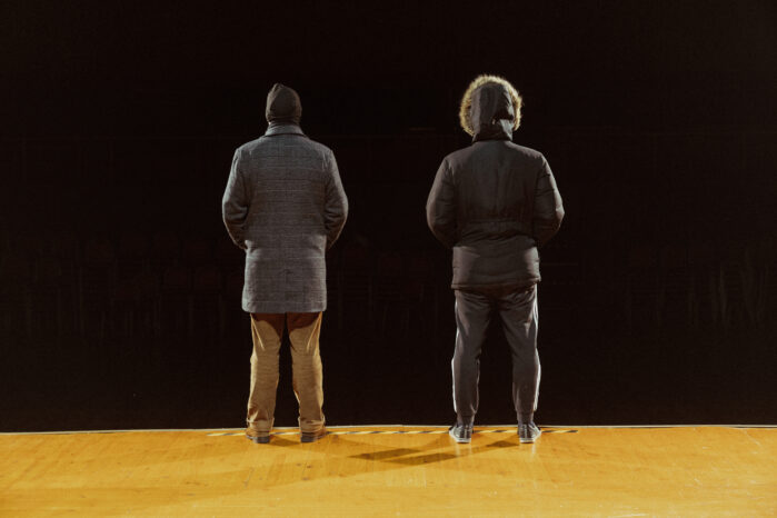 Kaksi henkilöä seisovat näyttämön reunalla selin kameraan.