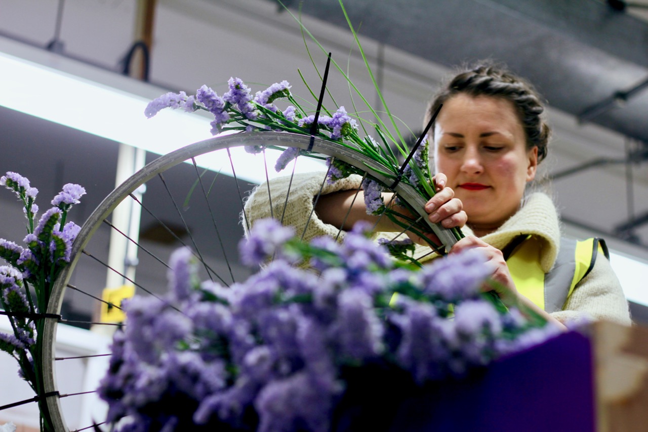 A dark haired woman is attaching flowers to a bicycle wheel.