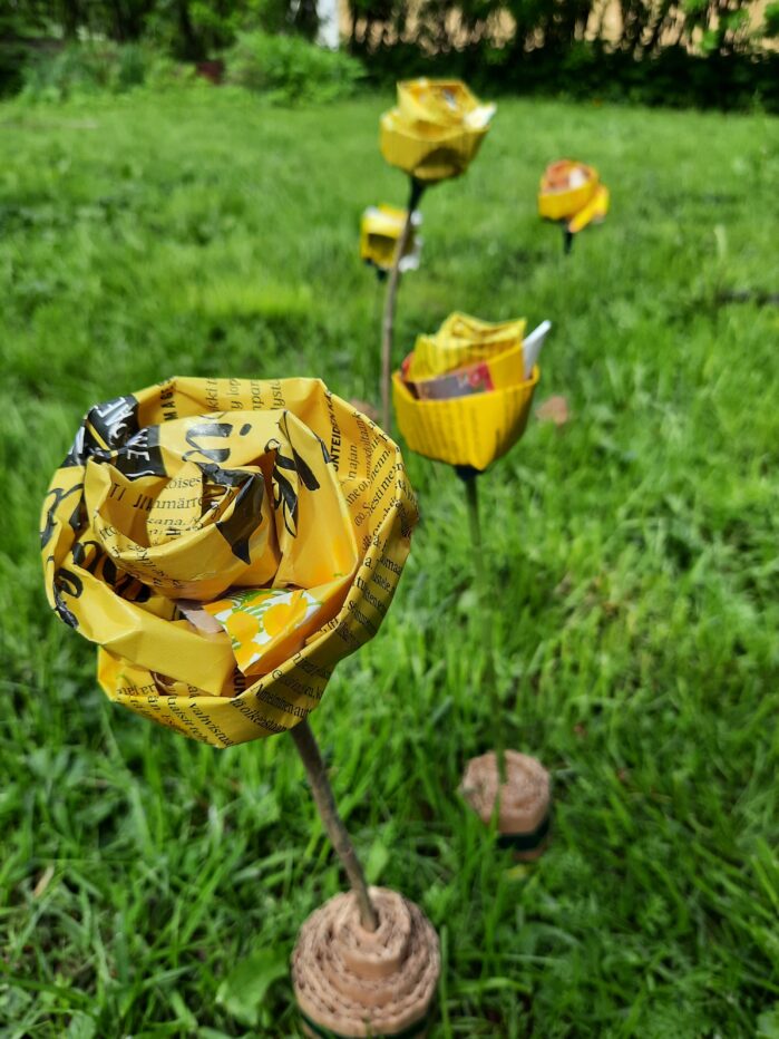 Vihreällä nurmella aikakausilehtien sivuista valmistettuja keltaisia kukkia, joiden varret ovat tehty luonnosta löytyneistä oksista.