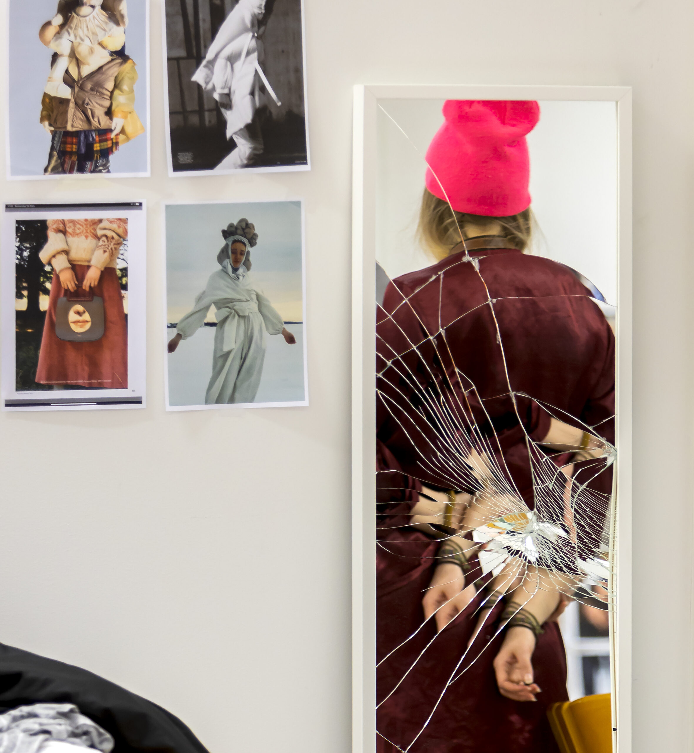 A broken mirror relfects a human figure. Pictures on the wall next to it.