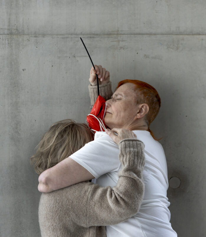Kaksi ihmistä halaa toistaan betoniseinän edessä. Toisen silmät ovat kiinni.