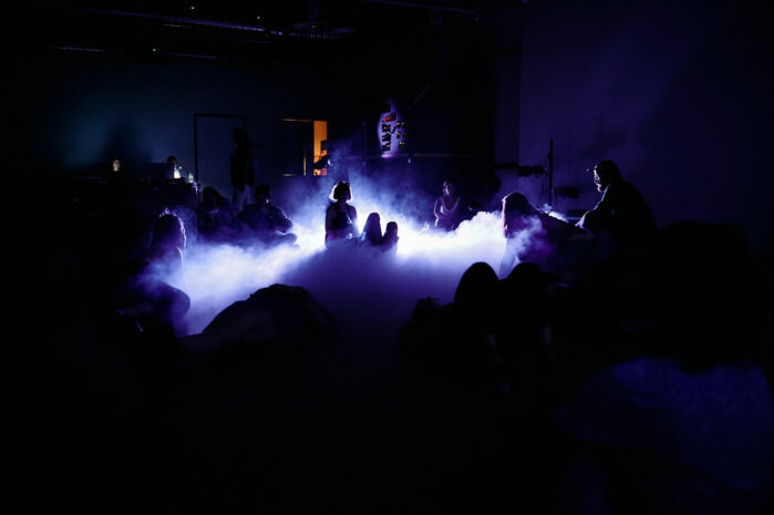 Dark room, shade of blue light in a mist. Some people in the room.