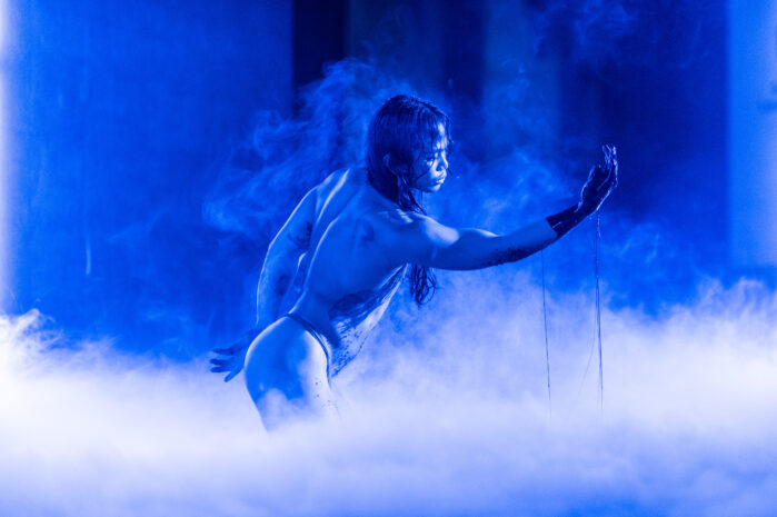 Blue light surrounds a person raising their hand. Performer has long hair and dark liquid covering their hand.