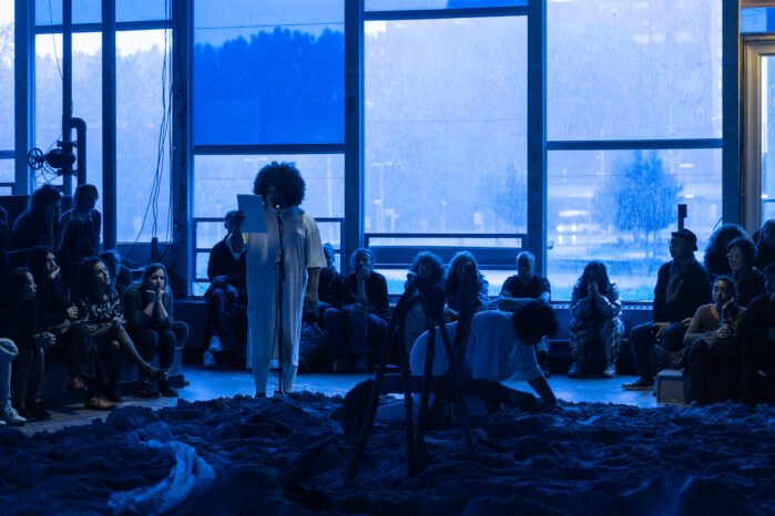 Blue lighted room where a group of people are watching Jota Mombaca and Tessa Mars performing. They are reading from a paper and there is sand in the floor.