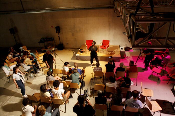 A crowd of people indoors sitting on chairs and looking at a performer. Two red chairs behind her.