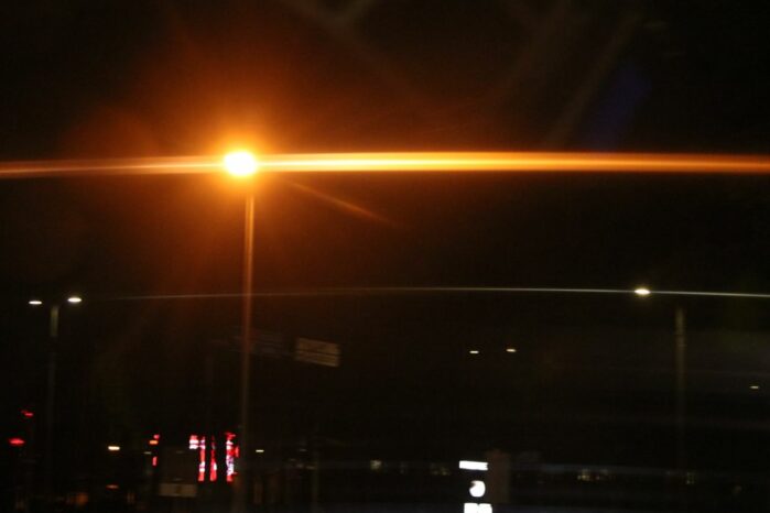 Öinen kaupunkimaisema. Tummasävyisen kuvan poikki kulkee kirkas, katuvalon aiheuttama valojuova.