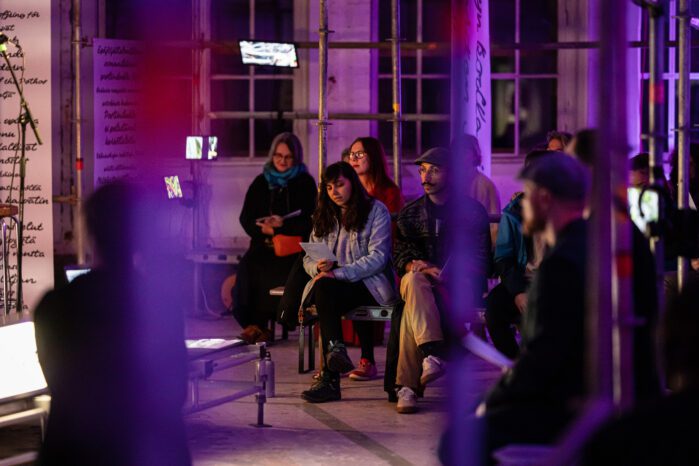 People sitting and watching an art performance in an industrial space. The space is lit with violet light.