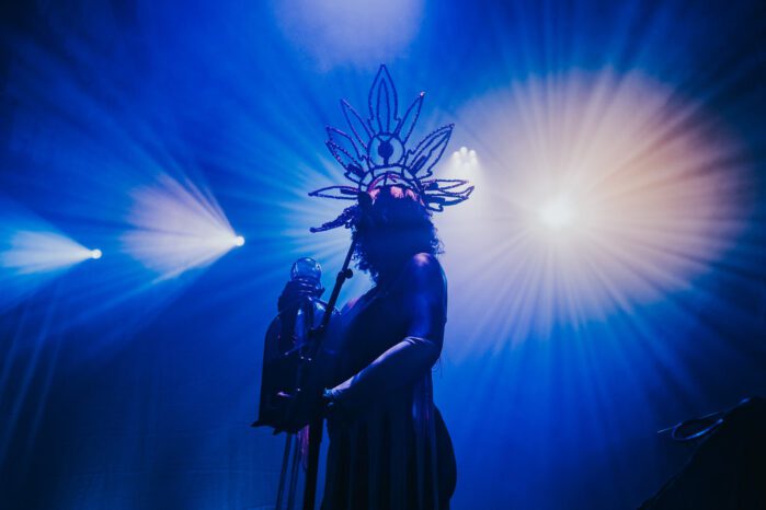 The silhouette of a person wearing a large tiara as they stand behind a microphone holding an object in their hands. The image is filled mostly with a deep blue light with bursts of white lights.