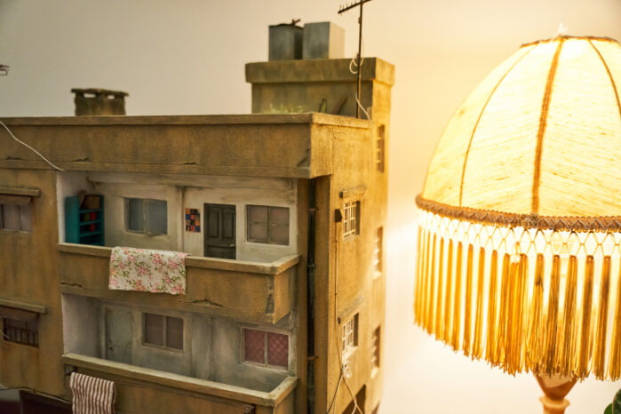 A miniature sculpture of a building with three floors and balconies, some fabrics hanging on the side of a balcony. Next to the sculpture is a lamp.