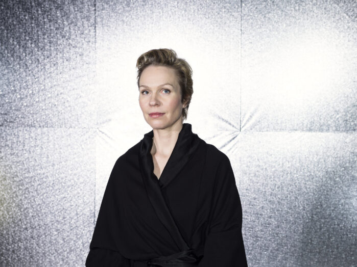 Maija Hirvanen wearing a black shirt, looking at the camera, infront of a light surface.