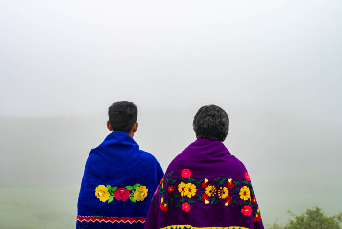 Kaksi ihmistä, nuorempi ja vanhempi, katsoo sumuista laaksoa kohti. Heillä on yllään tekstiilit, jotka on koristeltu värikkäillä kirjotuilla kukkasilla.