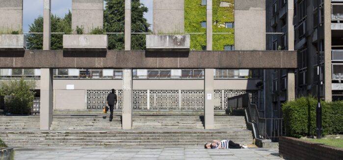 Outdoor urban landscape: Large concrete steps and columns, foliage and sky seen in the background. To the right someone is walking up the steps with their back to the camera. At the bottom right of the steps two people are lying down together, facing towards the camera, with their eyes closed.