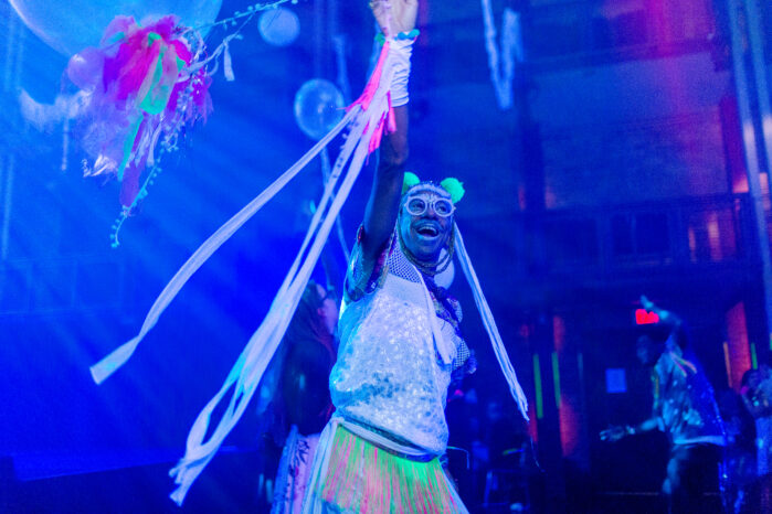 An image of a person on stage with one arm raised in the air. They are dressed in neon and light clothing with white streamers hanging from their raised arm. They have a joyful expression on their face with their mouth open in a smile. Behind them are bright club lights, white balloons and some people dancing.