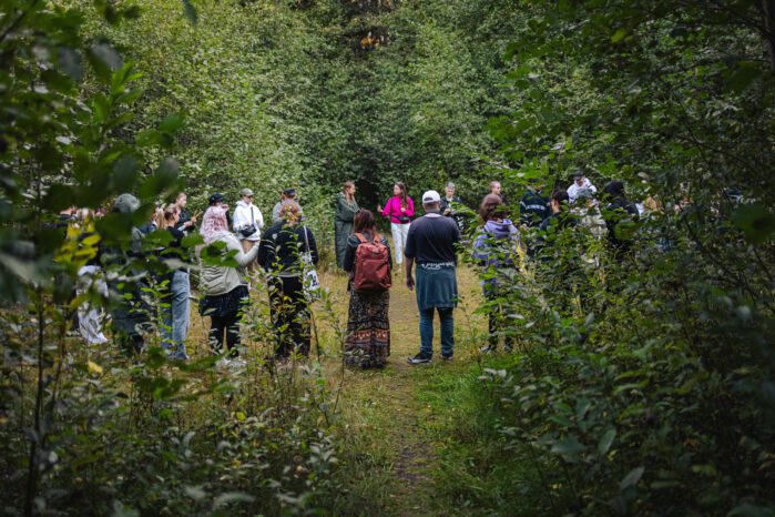 A group of people is gathered in circle. They are in the middle of a forest. It's a summer evening.