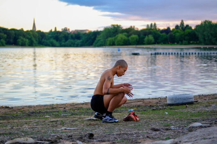 Sym, a transmasc lightskinned Black person, is crouching on a patch of sandy grass, in front of a cow heart. A setting sun casts a light on the body of water behind them. They are topless, showing visible tattoos on their arm, and have strings of black thread between their fingers that connect to the cow heart on the ground.