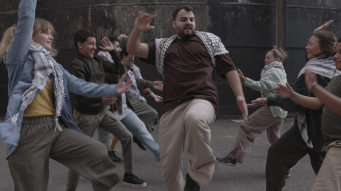 A diverse group of people forms a semicircle around a young man who dances and claps, with others clapping and dancing along.